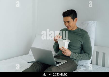 Ritratto di un uomo asiatico che mangia cibo da asporto guardando un computer portatile a letto. Nuovo stile di vita, ordine alimentare dal concetto di tecnologia moderna. Foto Stock