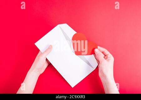 Una mano femminile apre una busta con un cuore rosso su uno sfondo rosso. Lettera d'amore al tuo amato. Dichiarazione d'amore. Invito al matrimonio. Visualizza da Foto Stock