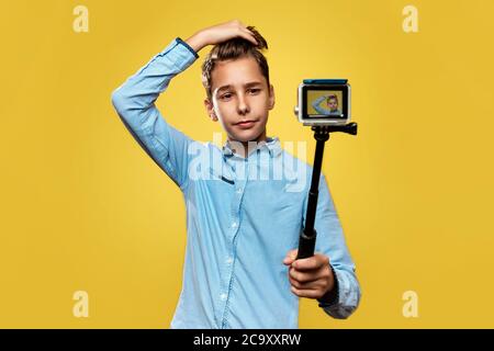 Bel blogger ragazzo fa un selfie o un video su una fotocamera compatta con il monopod. Sfondo giallo. Spazio di copia. Foto Stock