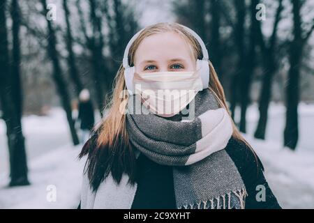 Giovane ragazza in maschera medica protettiva camminare nel parco invernale e ascoltare musica con le cuffie wireless Foto Stock