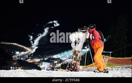 Coppia di turisti che si trovano sulla collina baciando sullo sfondo di notte stupefacente di piste illuminate da sci, copy space. Concetto di stile di vita attivo, sci notturno e relazioni. Foto Stock