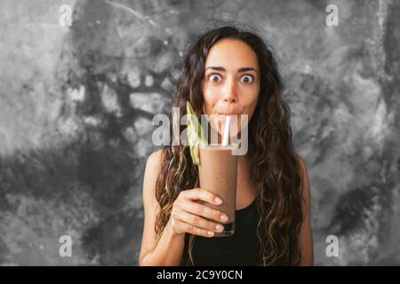 Divertente donna felice bere latte di cioccolato agitare con cacao e ridere. Concetto di bevanda estiva. Fondo parete in cemento grigio. Foto Stock