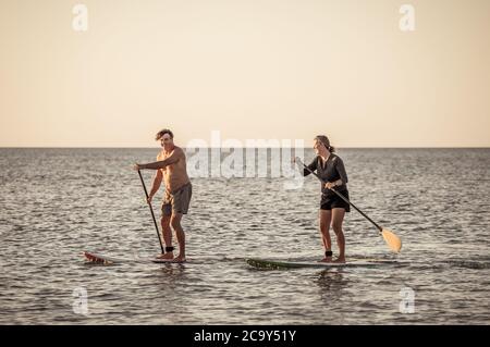 Coppia caucasica matura su SUP paddleboarding divertendosi su un mare tranquillo al tramonto in una bellissima spiaggia remota. Persone anziane moderne attive, all'aperto A. Foto Stock