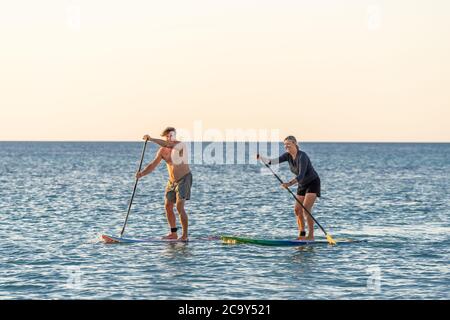 Coppia caucasica matura su SUP paddleboarding divertendosi su un mare tranquillo al tramonto in una bellissima spiaggia remota. Persone anziane moderne attive, all'aperto A. Foto Stock