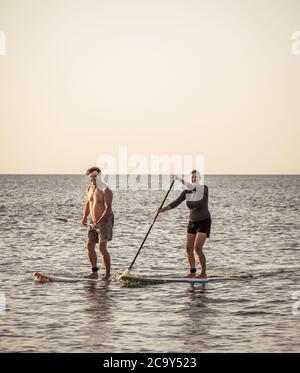 Coppia caucasica matura su SUP paddleboarding divertendosi su un mare tranquillo al tramonto in una bellissima spiaggia remota. Persone anziane moderne attive, all'aperto A. Foto Stock