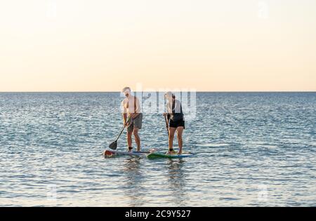 Coppia caucasica matura su SUP paddleboarding divertendosi su un mare tranquillo al tramonto in una bellissima spiaggia remota. Persone anziane moderne attive, all'aperto A. Foto Stock