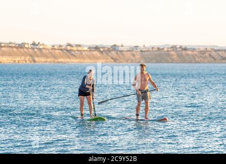 Coppia caucasica matura su SUP paddleboarding divertendosi su un mare tranquillo al tramonto in una bellissima spiaggia remota. Persone anziane moderne attive, all'aperto A. Foto Stock