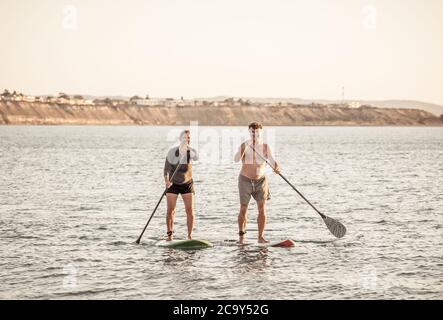 Coppia caucasica matura su SUP paddleboarding divertendosi su un mare tranquillo al tramonto in una bellissima spiaggia remota. Persone anziane moderne attive, all'aperto A. Foto Stock