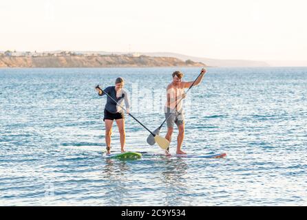 Coppia caucasica matura su SUP paddleboarding divertendosi su un mare tranquillo al tramonto in una bellissima spiaggia remota. Persone anziane moderne attive, all'aperto A. Foto Stock