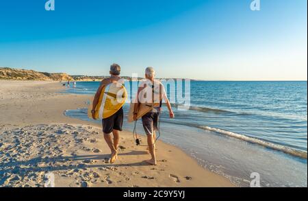 Due uomini maturi che camminano con tavole da surf sulla bellissima spiaggia godendo di paradiso e di vita di riposo. Amici adulti in forma attraente che si divertono Foto Stock