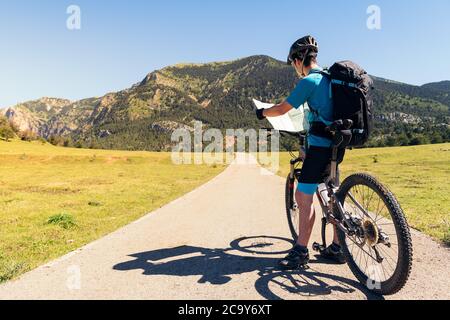 giovane ciclista con zaino e mountain bike in piedi su una strada mentre consulta il percorso della sua escursione sulla mappa, concetto di sport e sano Foto Stock