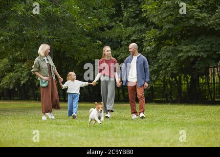 Ritratto grandangolare di famiglia spensierata con due bambini e cane da compagnia che tiene le mani mentre si cammina verso la macchina fotografica su erba verde all'aperto, spazio copia Foto Stock