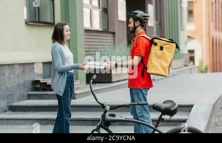 Online ordinazione di cibo a casa. Consegna uomo in casco con zaino giallo e bicikle dà scatole di pizza al cliente sulla strada, vicino casa Foto Stock