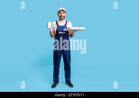 A lunghezza intera ha scioccato il corriere sorpreso in workwear e guanti di sicurezza che trasportano la scatola della pizza, mostrando il telefono mobile con il mock-up display per l'immagine di pubblicità. Foto Stock