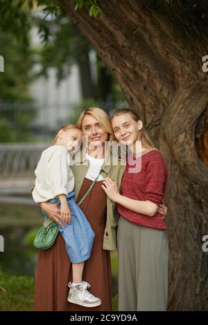 Ritratto verticale di madre moderna adulta con due figlie che si pongono insieme sorridendo felice guardando la macchina fotografica mentre si levano in piedi da albero all'aperto godendo il tempo della famiglia nel parco, copy space Foto Stock