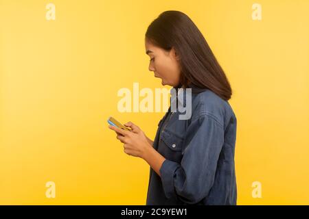 Vista laterale della ragazza sorpresa in elegante camicia denim digitando messaggio ed esprimendo shock, chiamando il numero di telefono cellulare, utilizzando on-line appli cellulare Foto Stock