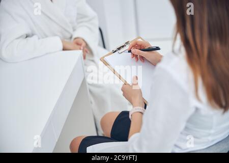 Dottore della clinica di bellezza che scrive qualcosa mentre si siede sulla sedia Foto Stock