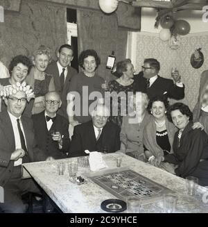 1960, storico, festa di compleanno per adulti in una casa, uomini e donne ben vestiti che festeggiano con alcune bevande, gin e un gioco di roulette da tavolo, con una piccola ruota e tavola seduti su un tavolo coperto di stoffa sala da pranzo, Inghilterra, Regno Unito. Foto Stock