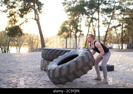 Allenamento uomo forte sollevamento pneumatico grande palestra fai da te all'aperto. Sportivo giovane adulto ragazzo caucasico girare grande ruota sulla natura. Attrezzature sportive artigianali Foto Stock