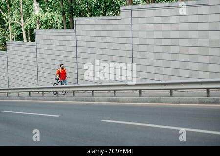 Percorso di consegna. Corriere in casco, con zaino giallo e passeggiate in bicicletta lungo la pista in strada per il cliente Foto Stock