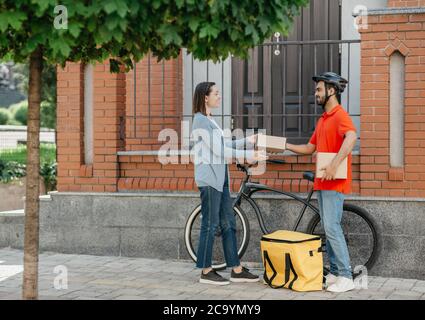 Acquisto in negozio online e consegna tramite corriere. La ragazza sorridente riceve il pacco dal deliveryman con la bicicletta sulla strada Foto Stock