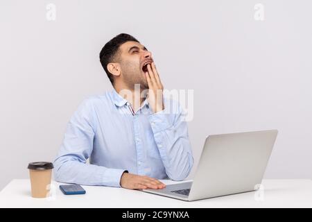 Annoiato un impiegato uomo esausto seduto in ufficio con un computer portatile sulla scrivania, sbadigliare il viso con la mano, sensazione di stanchezza di lavoro straordinario, notte s Foto Stock