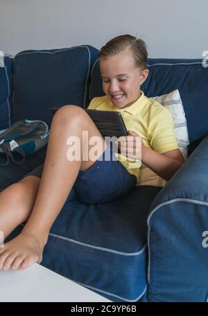 Ragazzo giovane positivo in abiti casual seduto sul divano e utilizzando tablet mentre fare i compiti a casa. Foto Stock