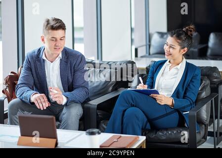 Sorridente giovane donna d'affari che prende appunti nel pianificatore quando parla con un collega che mostra la presentazione sul computer tablet Foto Stock