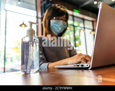 Giovane donna asiatica che usa un computer portatile che lavora a casa con gel di alcol in quarantena per coronavirus indossare maschera protettiva con distanza sociale Foto Stock