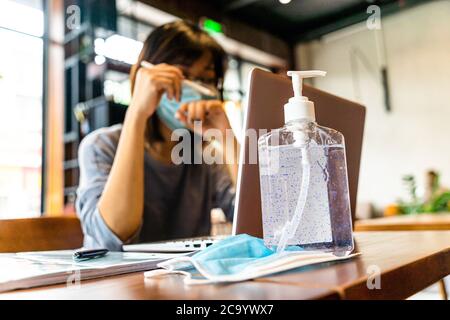 Giovane donna asiatica che usa un computer portatile che lavora a casa con gel di alcol in quarantena per coronavirus indossare maschera protettiva con distanza sociale Foto Stock