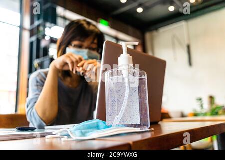 Giovane donna asiatica che usa un computer portatile che lavora a casa con gel di alcol in quarantena per coronavirus indossare maschera protettiva con distanza sociale Foto Stock