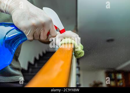 Giovane uomo asiatico in guanti protettivi disinfezione ringhiere scale durante la pulizia a casa, vista ravvicinata sulle mani e con flacone spray. Assistenza sanitaria e. Foto Stock