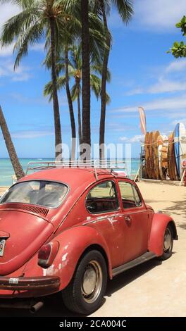 Un betle rosso VW sulla spiaggia di Waikiki con palme e tavole da surf contro un cielo blu Foto Stock