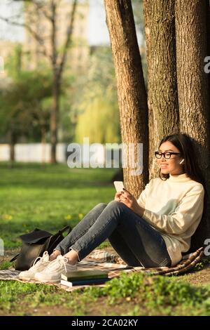 Studentessa di sesso femminile che usa il telefono, seduto sotto l'albero nel parco Foto Stock