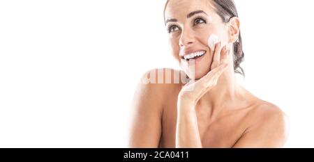 Bella giovane donna che applica crema al suo viso isolato su bianco. Concetto di benessere e cosmetici per la cura della pelle. Foto Stock
