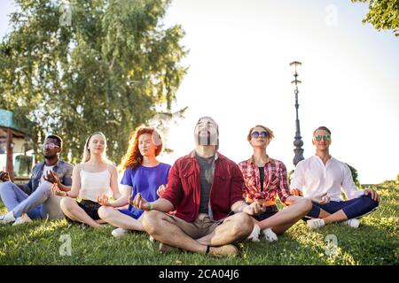 Diversi amici caucasici e afro-americani adulti che amano rilassarsi yoga all'aperto nel parco. Praticare yoga in paesaggio naturale, con l'uomo guardato sopra Foto Stock