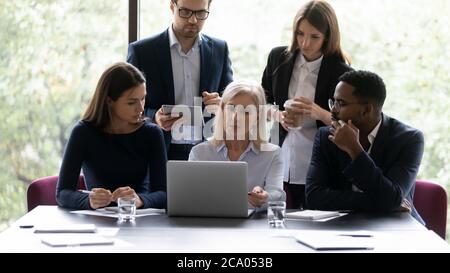 Mentore di mezza età insegnano ai giovani dipendenti multietnici utilizzando un'applicazione per computer Foto Stock