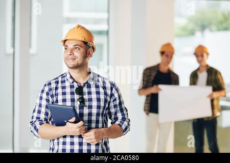 Sorridendo giovane ingegnere in hardhat che tiene un tablet e guardando all'interno della casa il suo team ha appena finito Foto Stock