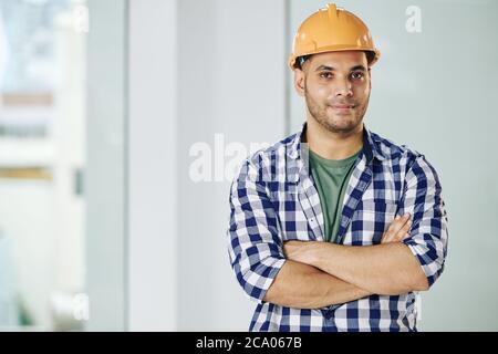 Ritratto di bel contrattista sorridente in hardhat pieghevole braccia e guardando la macchina fotografica Foto Stock