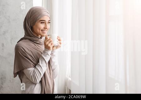 Rilassante donna araba a Hijab gustando il caffè mattutino vicino alla finestra a casa Foto Stock