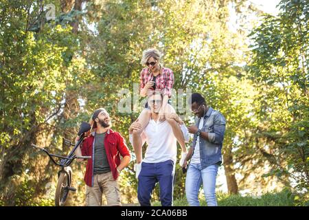 Giovane pilota BMX in compagnia dei suoi amici multietnici che camminano per divertire al parco cittadino, felice giovane donna è piggybacking sul suo ragazzo Foto Stock