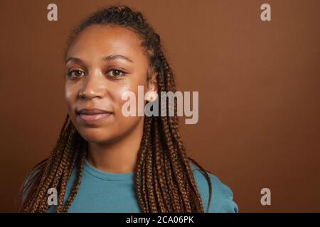 Ritratto minimo di testa e spalle di una moderna donna afroamericana che sorride con la fotocamera mentre si sta in piedi contro sfondo marrone in studio, spazio di copia Foto Stock