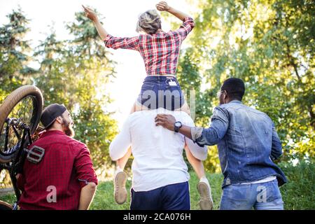 Vista posteriore di persone multirazziali non riconosciute che andavano al parco nel pomeriggio estivo. Ragazzo che porta la sua fidanzata sul piggyback e uno dei ragazzi auto Foto Stock