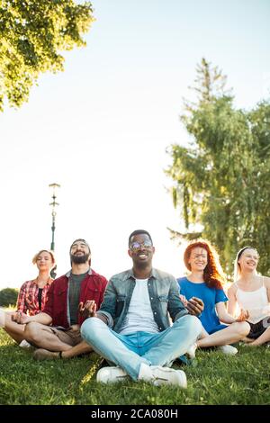 Giovane afroamericano che indossa occhiali da sole, che conduce una lezione di yoga in un parco pubblico all'aperto. Amici multirazziali in abiti estivi casual decidere di ha Foto Stock