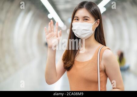 Giovane donna asiatica passeggero che indossa una maschera chirurgica e guardando la macchina fotografica per ciao o salutare con il suo amico in interscambio metropolitana quando si viaggia in b Foto Stock
