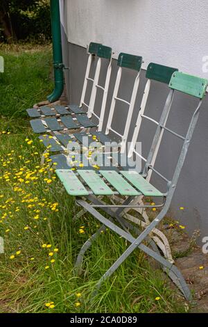 Quattro vecchie sedie verdi sbiadite da giardino di fronte ad un muro con erba e fiori che crescono intorno a loro Foto Stock