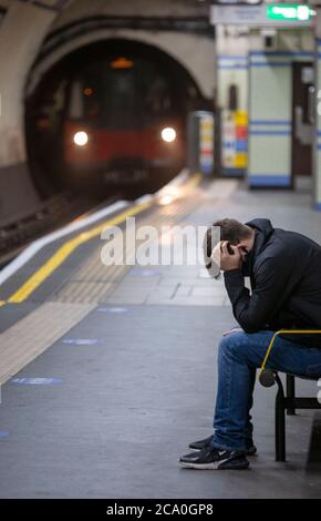 Il giovane si siede con la testa nelle mani su una stazione della metropolitana di Londra, con il treno in avvicinamento. Londra, Inghilterra Regno Unito Foto Stock