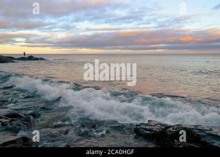 Le nuvole dalle sfumature di rosa si sollevano dietro una figura umana lontana in piedi Rocce che si aggettano sul lago Superior Foto Stock