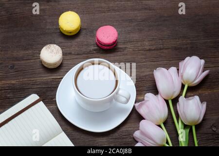 Vista dall'alto su una tazza di tè, macaron, tulipani rosa e notebook. Sfondo di legno. Foto Stock