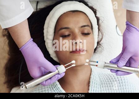 Giovane donna felice sulle procedure cosmetiche moderne. Concetto di spa per la cura della pelle. Closeup di bella donna che riceve trattamento viso microcorrente da terapeuta a Spa Salon. Foto Stock